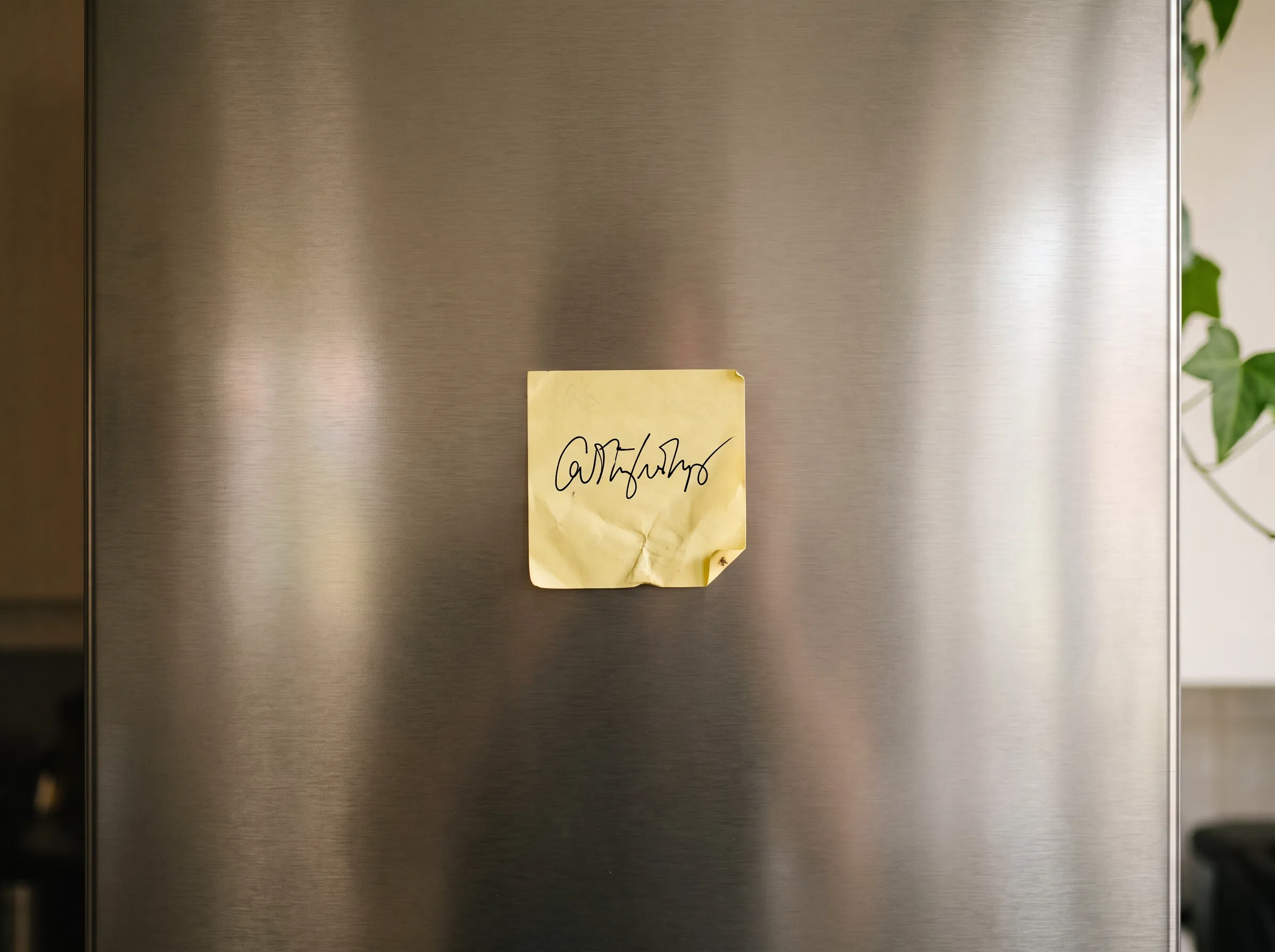 Ein einzelner gelber Post-it-Zettel mit einer kurzen handschriftlichen Notiz an einem Kühlschrank aus Edelstahl